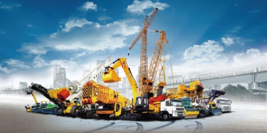 A variety of yellow and white construction vehicles and heavy machinery are arranged in a semi-circle on a paved surface. The equipment includes cranes, excavators, trucks, and loaders. In the background, there is a cityscape with tall buildings and a bridge under a partly cloudy blue sky.