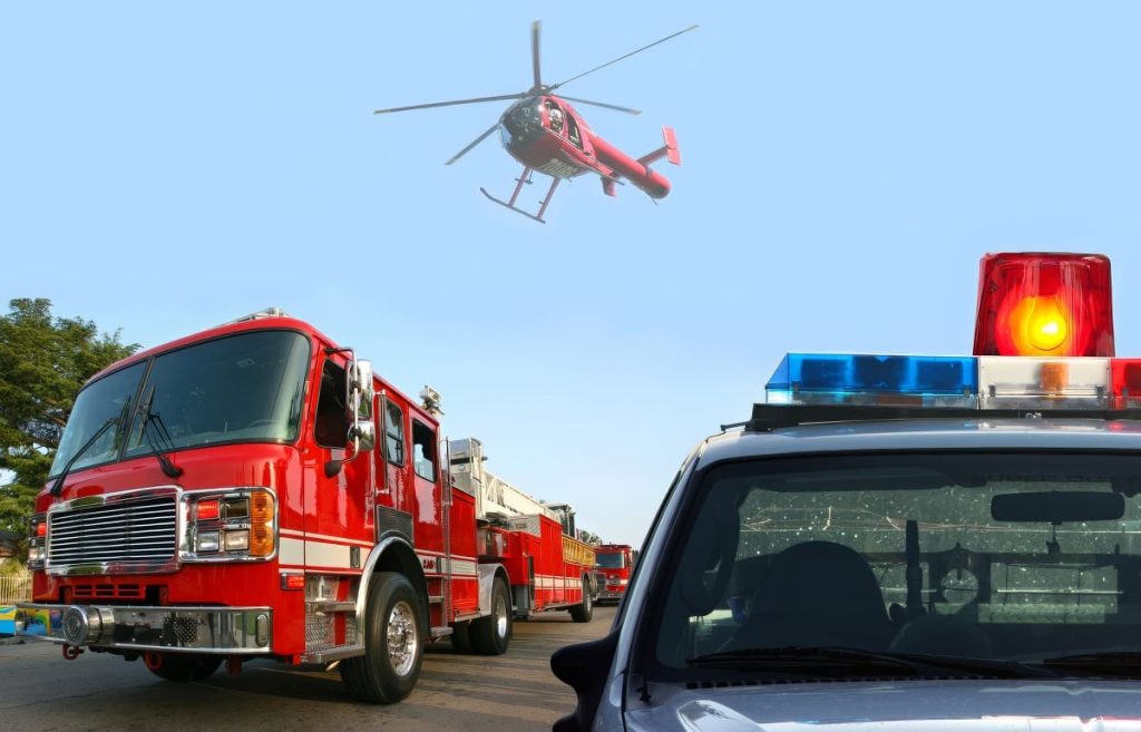 Red fire truck parked on a street, a police car with flashing red and blue lights in the foreground, and a red helicopter flying in the clear blue sky above.