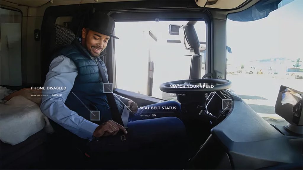 A man wearing a black cap, blue shirt, and dark vest is seated inside a vehicle cabin, fastening his seat belt. The cabin features a steering wheel and dashboard, with a side mirror visible on the right. Text overlays indicate "PHONE DISABLED," "SEAT BELT STATUS," and "WATCH YOUR SPEED," suggesting a focus on safety and vehicle status.